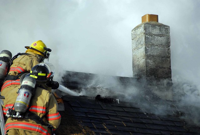 Two firefighters atop a burning residential roof producing large clouds of black smoke.